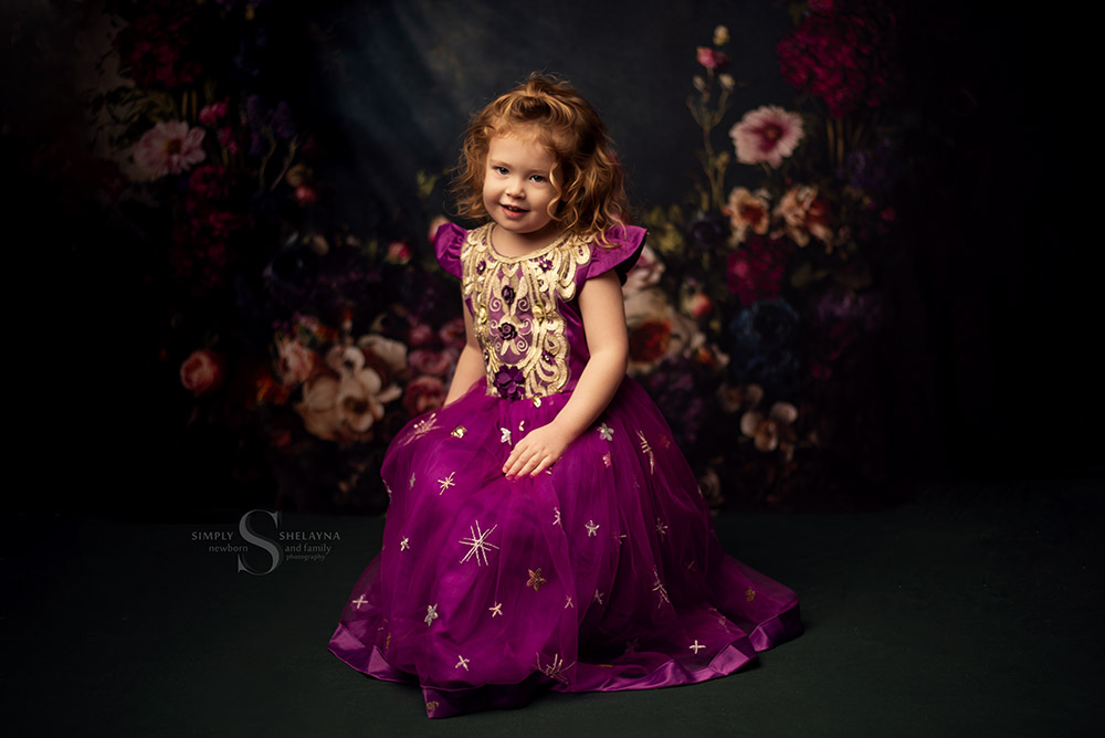 A toddler girl poses in front of a deep green flowered Kate Backdrop with Simply Shelayna Children's Photography in Syracuse NY.
