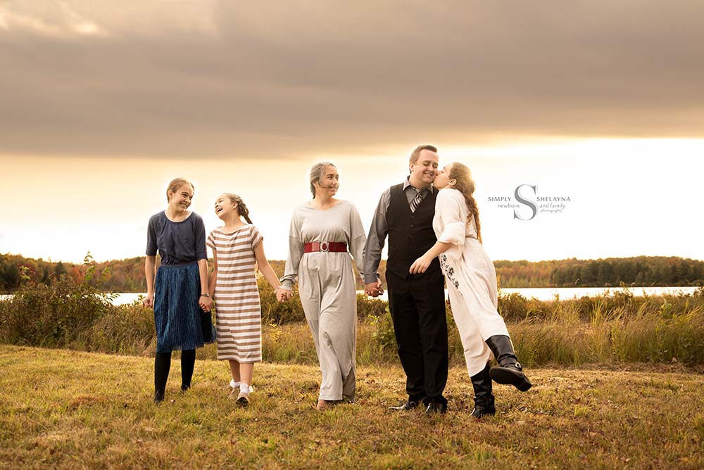 A family of 5 walk together holding hands as a daughter kisses her father with Simply Shelayna Family Photographer in Syracuse NY.