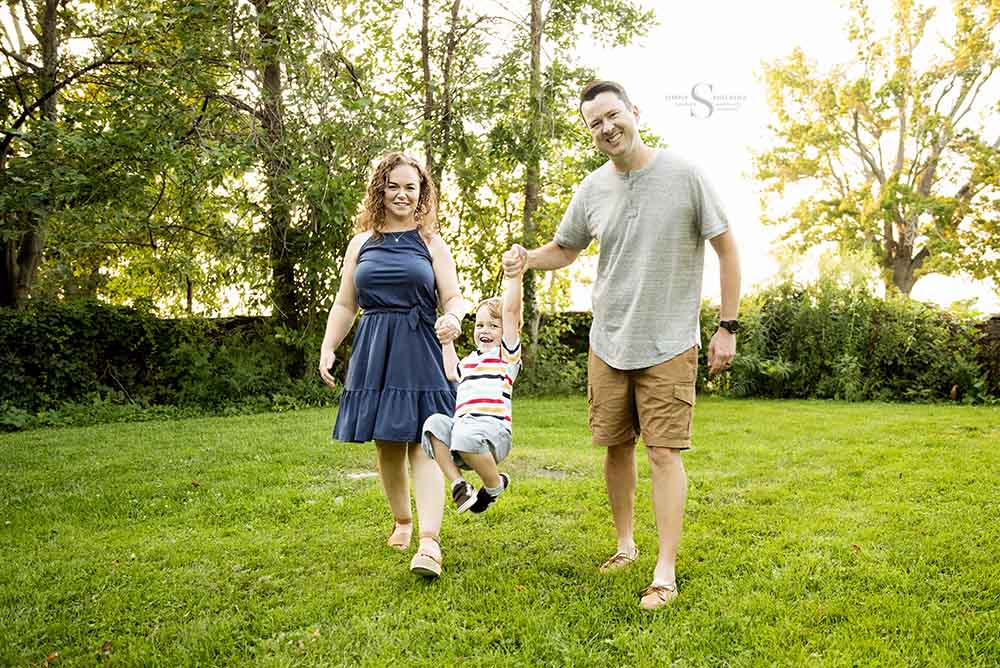 A mother and father swing their young son at the beautiful grounds of Mexico Point State Park for a summer evening portrait session with Simply Shelayna Family Photography in Oswego NY.