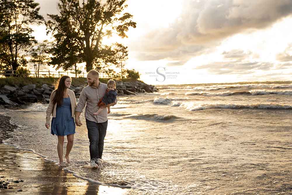 A family of 3 walks through the beach waters of Mexico Point State Park in Oswego County during a late summer sunset with Simply Shelayna Family Photography.