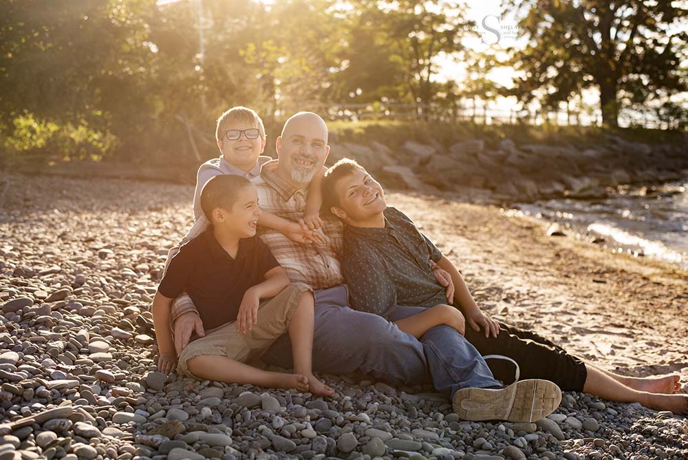 A father sits on a rocky beach surrounded by his 3 sons as they view Lake Ontario at Mexico Point State Park in CNY with Simply Shelayna Family Photography.