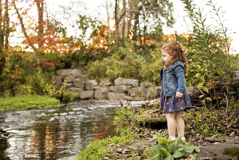 A toddler girl with red hair wearing a jean jacket, throws a stone into the brook at Fallbrook SUNY Oswego with Simply Shelayna Family Photography in Oswego NY.