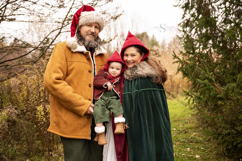 A family of three, dressed in red hand knitted pixie hats stroll through a wooded territory with baby sitting on an antique sled for family portraits in Syracuse NY with Simply Shelayna Family Photographer.