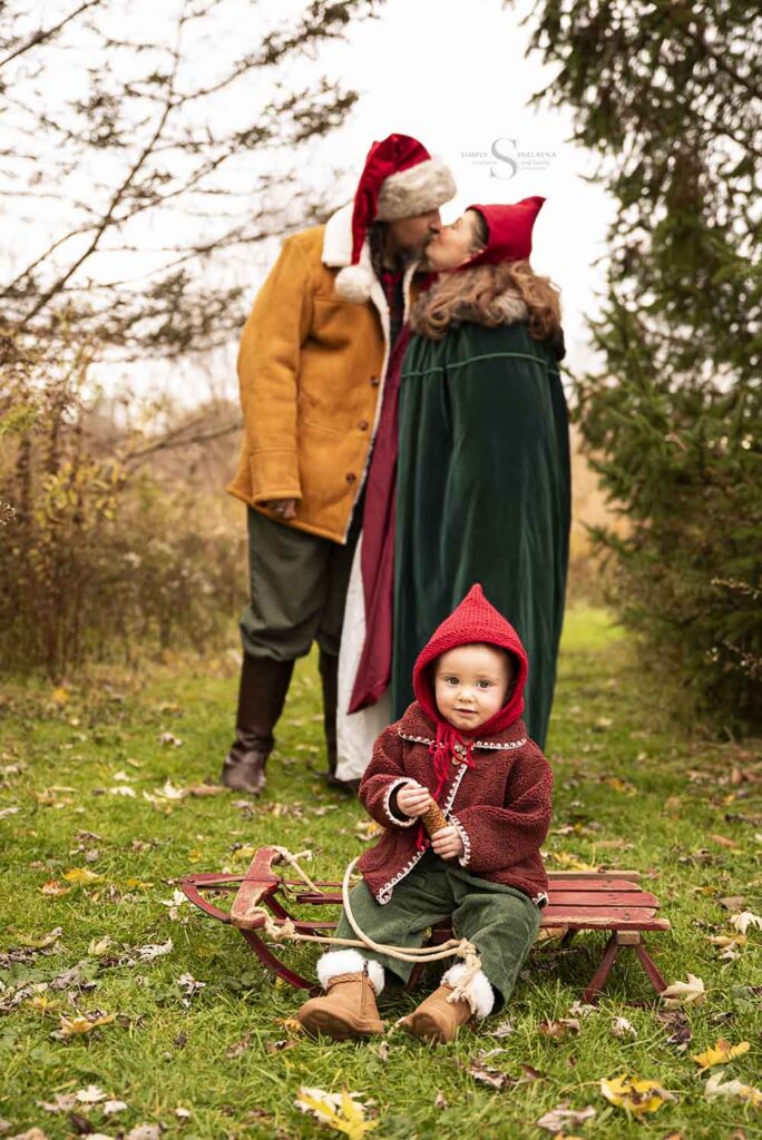 A family of three, dressed in red hand knitted pixie hats stroll through a wooded territory with baby sitting on an antique sled for family portraits in Syracuse NY with Simply Shelayna Family Photographer.