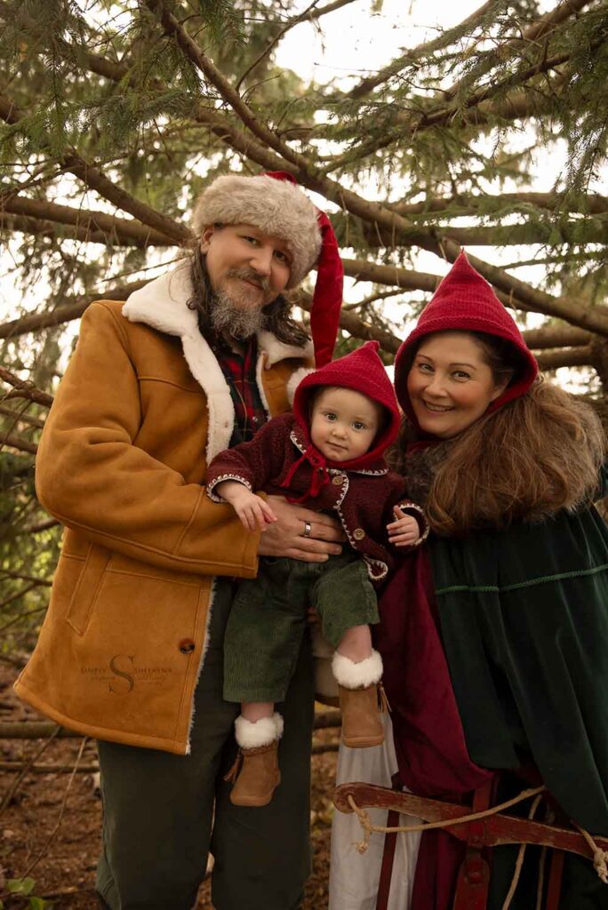 A family of three, dressed in red hand knitted pixie hats stroll through a wooded territory with baby for family portraits in Syracuse NY with Simply Shelayna Family Photographer.