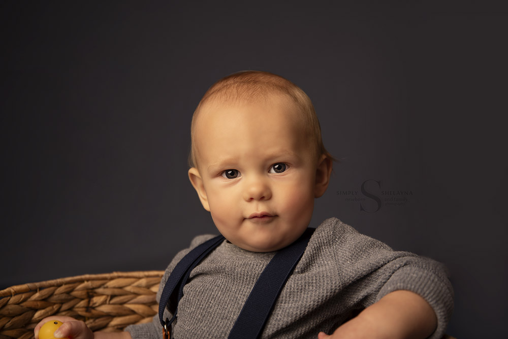 A baby boy sits up in a wicker fish shaped basket  for his nine month milestone portrait with Simply Shelayna Baby Photography in Oswego NY.