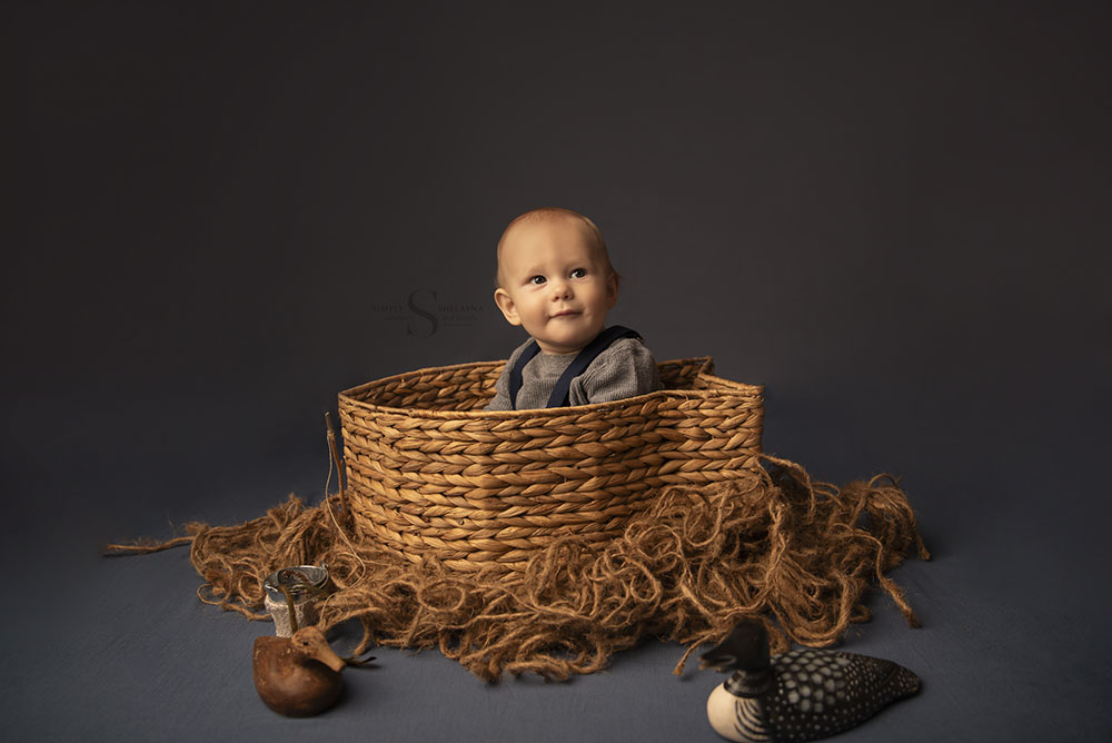 A baby boy sits up in a wicker fish shaped basket surrounded by woodland ducks for his nine month milestone portrait with Simply Shelayna Baby Photography in Oswego NY.