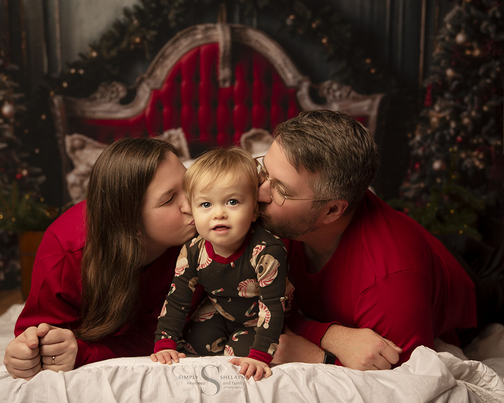 A family of 3, dressed in coordinating Christmas pjs, pose for a Night Before Christmas portrait with Simply Shelayna family photographer in Oswego NY.