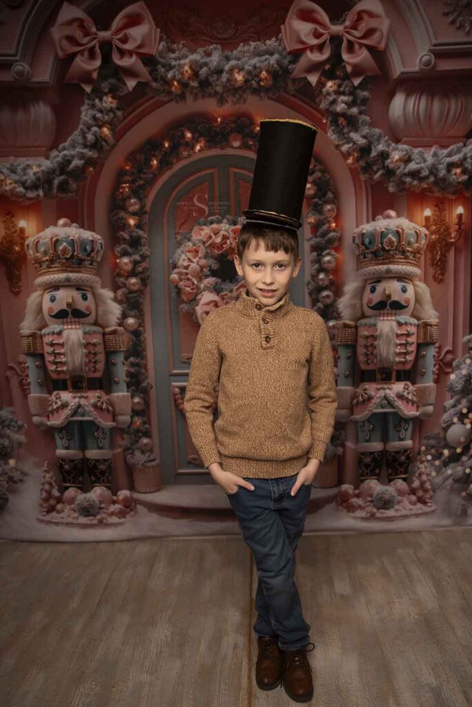 A young boy stands erect with a nutcracker hat tall upon his head for a Christmas portrait with Simply Shelayna Family Photography in Oswego NY.