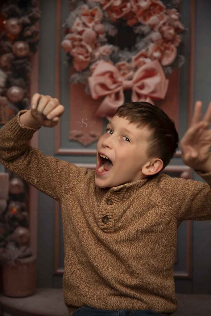 A young boy puts on a silly face while raising his hands during Christmas portraits with Simply Shelayna family photography in CNY.