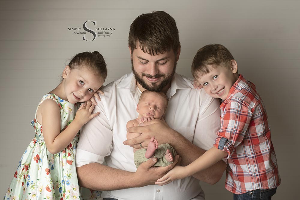 A father holds his newborn son, while is two young children hold onto him for a father and children portrait with Simply Shelayna Newborn Photography in Syracuse NY.