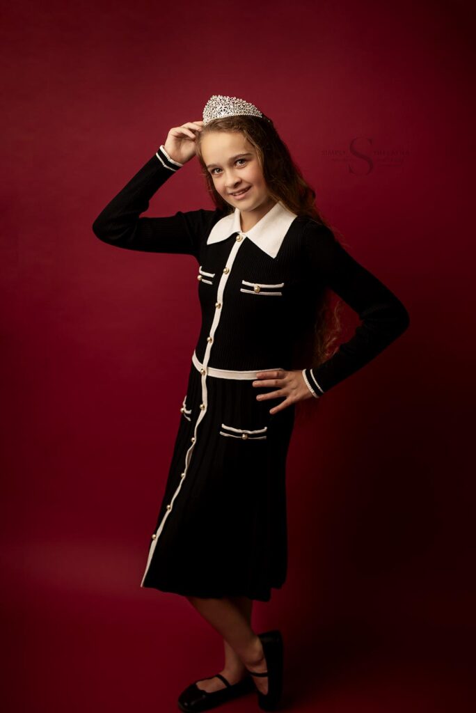 A pre-teen girl dressed in a black and white gown, posed on a red backdrop and wearing a princess tiara, poses for her birthday portraits with Simply Shelayna Family Photography in Syracuse NY. 