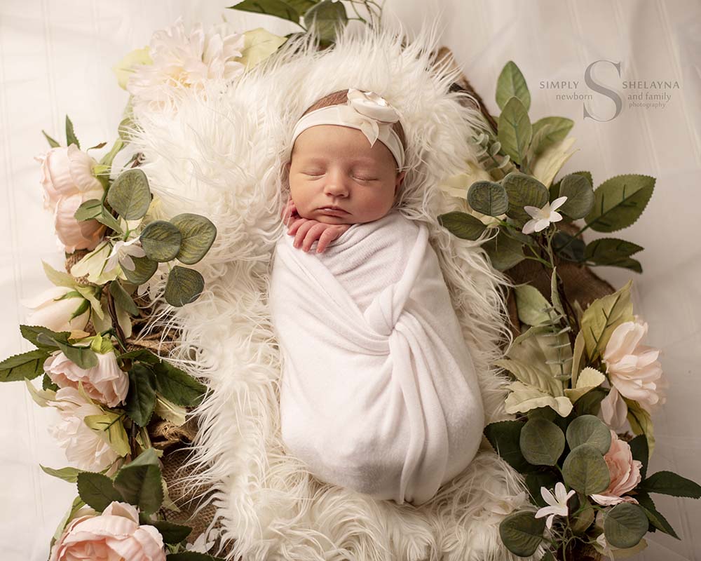 A newborn baby girl sleeps in a white flokati surrounded by flowers and greenery with Simply Shelayna baby photography in Syracuse NY. 