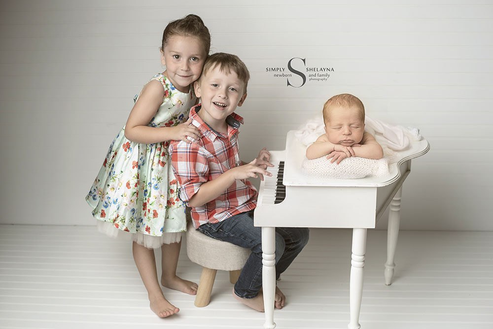 A newborn baby boy is posed chin up on a baby grand piano while is older siblings play the piano with Simply Shelayna Photography in CNY.
