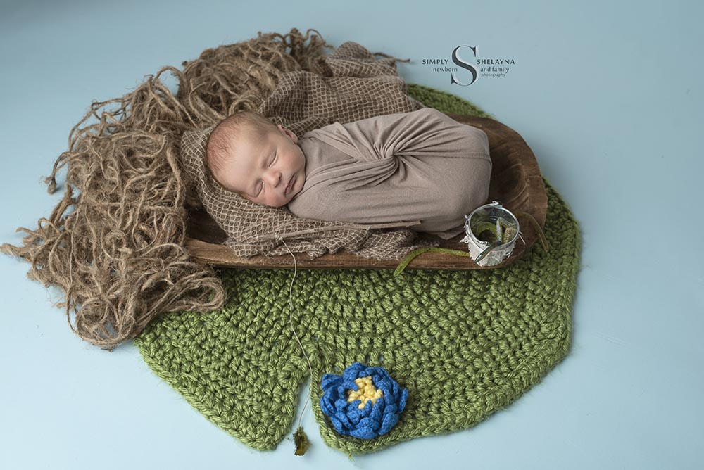 A baby boy is swaddled in a brown wrap and sleeps in a little boat upon a hand knitted lily pad with Simply Shelayna Newborn Photography in CNY.