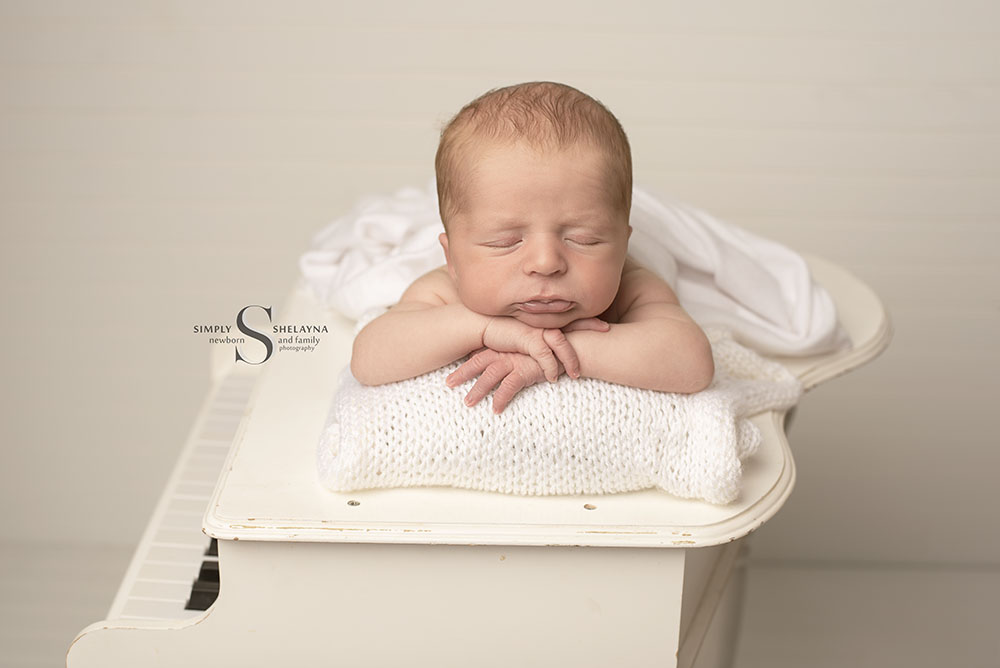 A newborn baby boy is posed chin up on a baby grand piano with Simply Shelayna Photography in CNY.