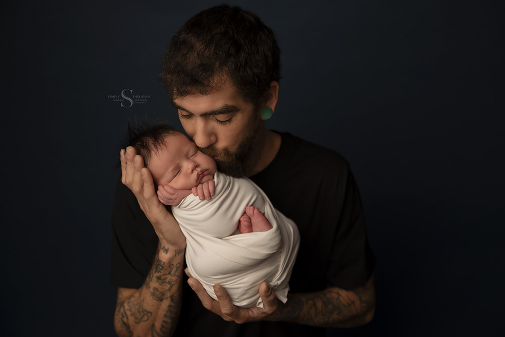 A father kisses his son who is swaddled in a white wrap for a portrait with Simply Shelayna Newborn Photography in Oswego NY.