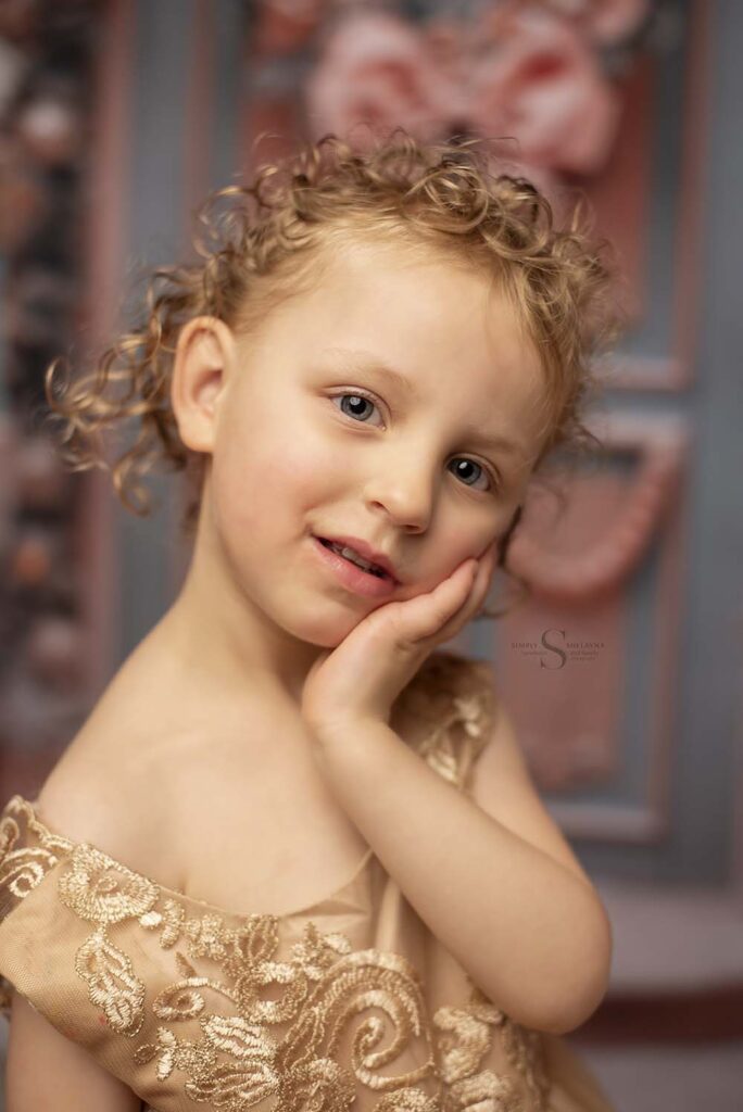 A young girl wears a gold Christmas dress, and holds her cheek in her hand for a portrait with Simply Shelayna Family Photography in Albany NY.