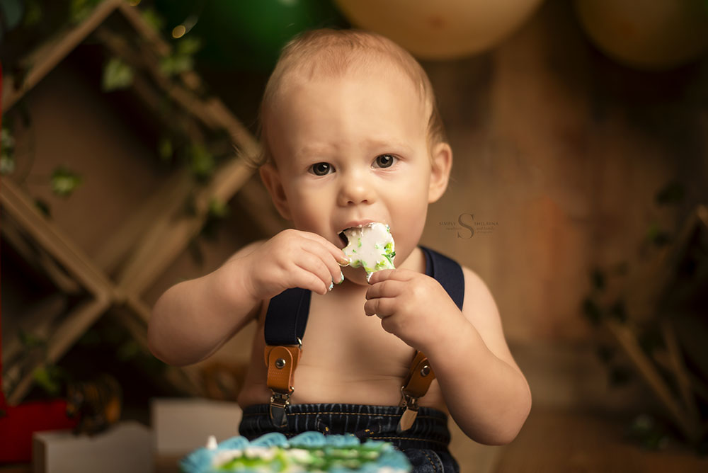 A 1 year old boy dressed in jeans and overalls enjoys his smash cake with Simply Shelayna Baby photography in Oswego NY.