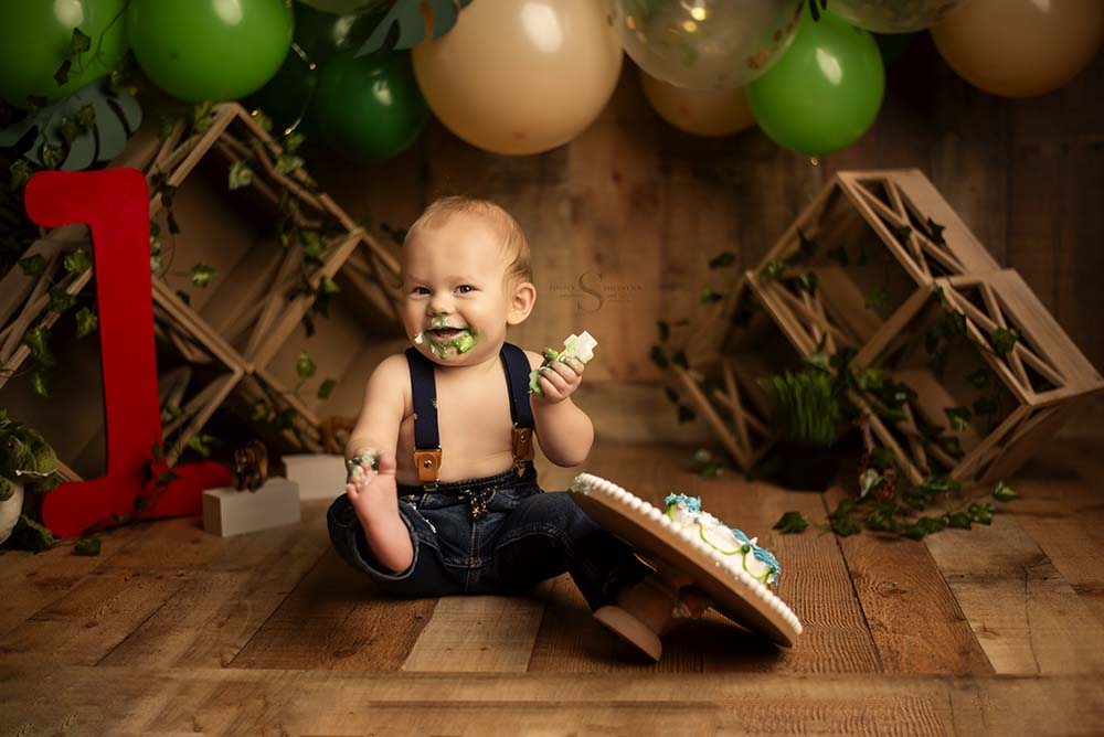 A 1 year old boy dressed in jeans and overalls tips over his smash cake with Simply Shelayna Baby photography in Oswego NY.