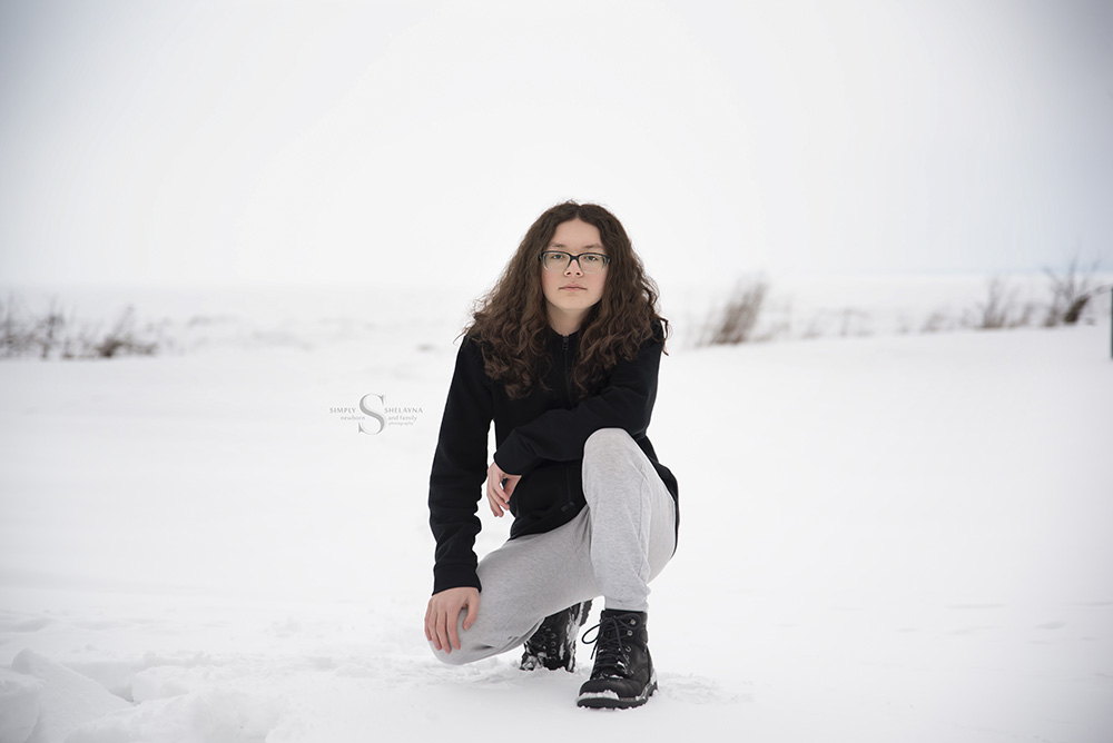 A young man crouches in the snow with a frozen Lake Ontario behind him for senior portrait with Simply Shelayna Photography.