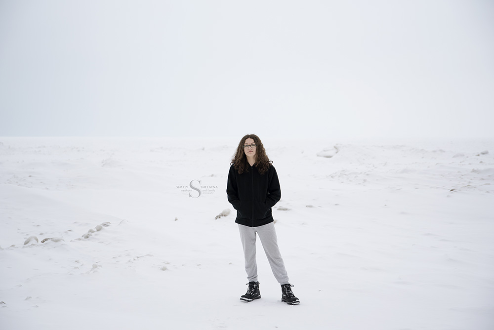 A nearly mono chromatic portrait of a young man standing in a snow white scene with a frozen Lake Ontario behind him with Simply Shelayna Senior Photography in Syracuse NY. 