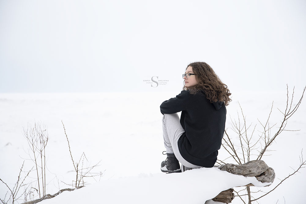 An highschool senior sits with his back to the camera in a snow white scene looking out to Lake Ontario at Mexico Point State Park with Simply Shelayna Family Photography.