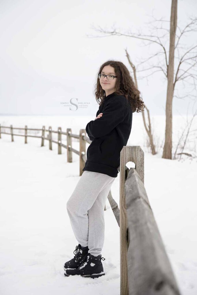 An high school senior stands up against a wooden fence with Lake Ontario beyond him, for a portrait with Simply Shelayna Senior photography in  CNY.