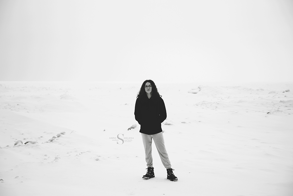 A mono chromatic portrait of a young man standing in a snow white scene with a frozen Lake Ontario behind him with Simply Shelayna Senior Photography in Syracuse NY. 
