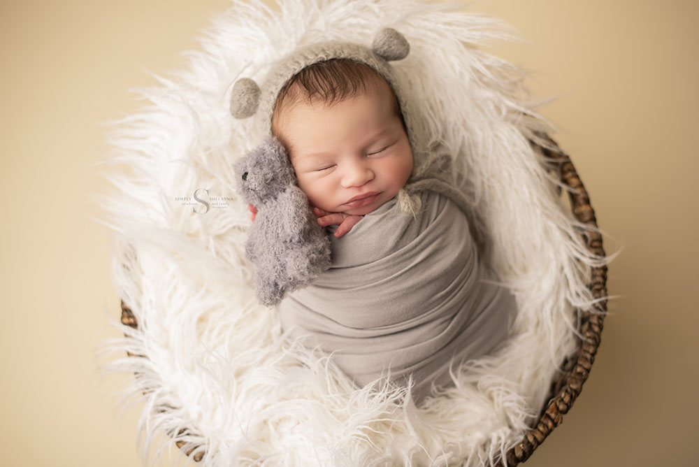 A newborn boy sleeps on a large white blanket placed in basket while snuggling a teddy bear and wearing a gray bear bonnet with Simply Shelayna Newborn Photography in Syracuse NY.