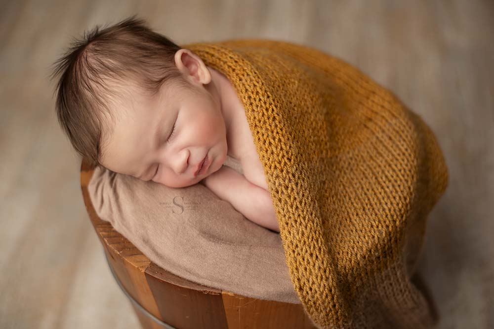 A newborn boy sleeps in a bucket with a knitted gold wrap draped over him with Simply Shelayna Newborn Photography in CNY.