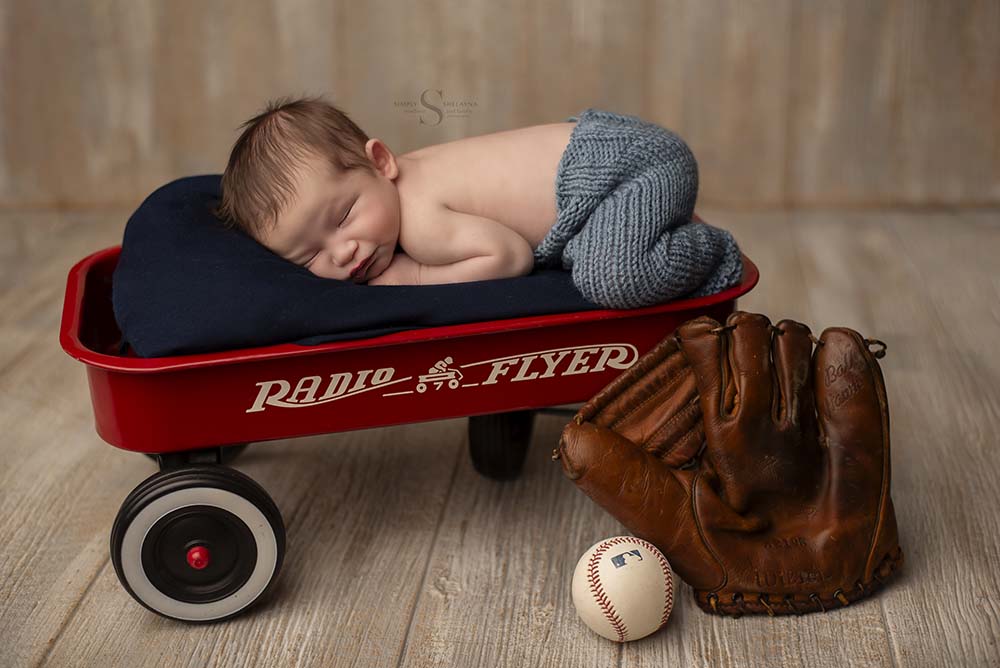 A newborn boy sleeps in a red rider wagon with a antique catcher's mitt and baseball in the picture for a newborn baseball themed portrait with Simply Shelayna Newborn photography in CNY.