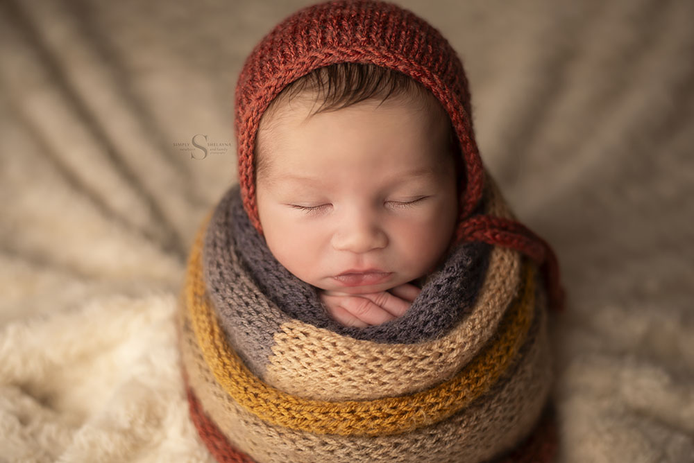 A sweet baby boy is wrapped in a handknitted multi color autumn wrap with a coordinating bonnet for a newborn portrait with Simply Shelayna Newborn Photography in Syracuse NY.