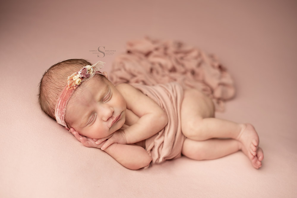 A baby girl sleeps on a soft pink backdrop for a newborn portrait with Simply Shelayna Newborn Photography in Oswego NY.