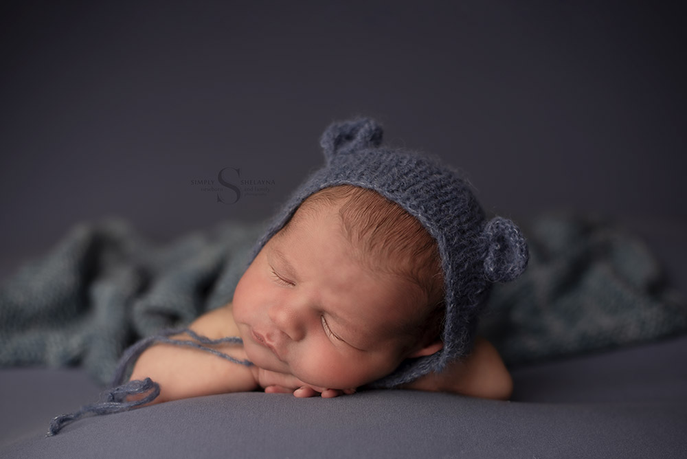 A baby boy sleeps with his chin on hands wearing a hand knitted blue bear bonnet with Simply Shelayna Newborn Photographer in Syracuse NY.