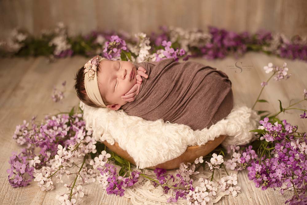 A newborn baby girl sleeps in a basket while surrounded by purple and white wildflowers with Simply Shelayna Newborn Photography in Oswego NY.