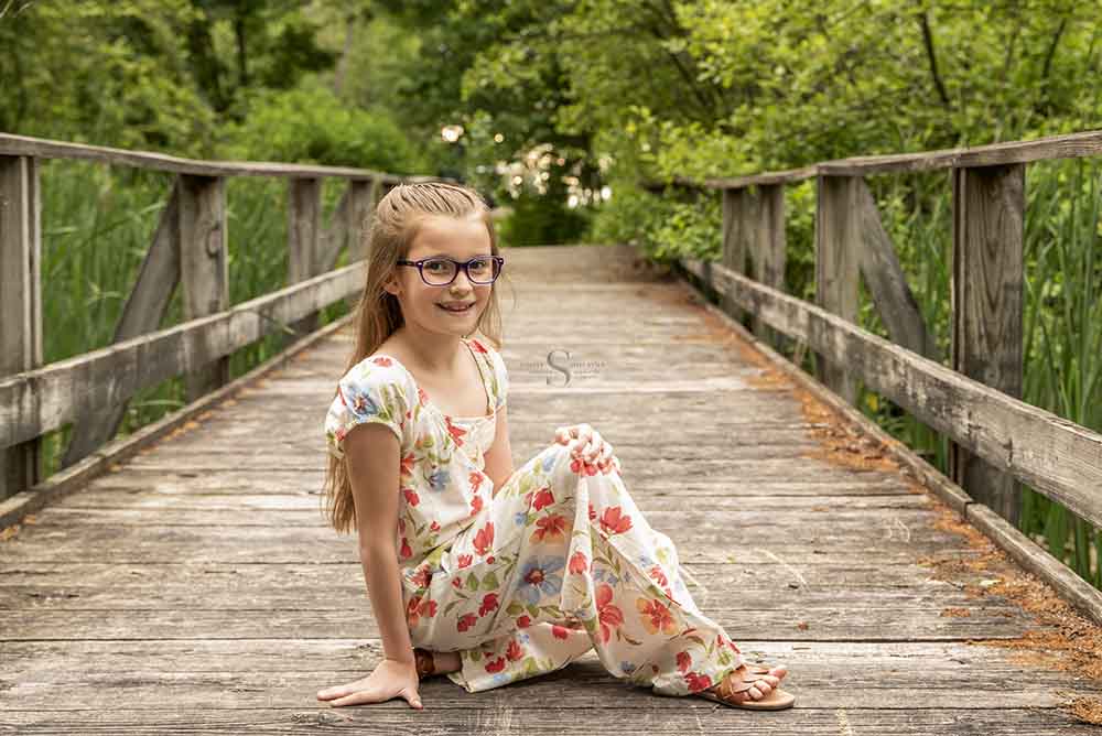 A young girl in a floral jumper sits posed on a boardwalk in Mexico Point State Park with Simply Shelayna Family Photography in Oswego NY.