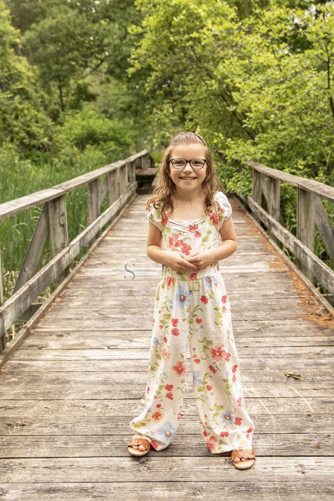 A young girl in a floral jumper poses on a boardwalk bridge with Simply Shelayna Family photography in Syracuse NY.