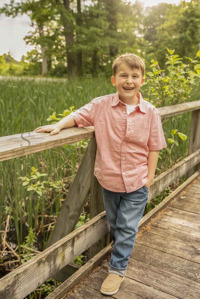 A young boy relaxes on a board walk railing for a portrait in Mexico Point State Park with Simply Shelayna Family Photography in Syracuse NY.