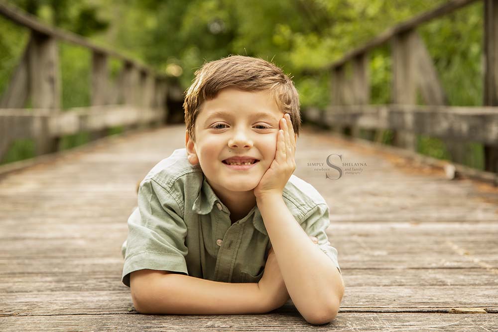 A young boy lies on his stomach with one hand under his cheek for a portrait on a boardwalk with Simply Shelayna Family Photography in CNY.