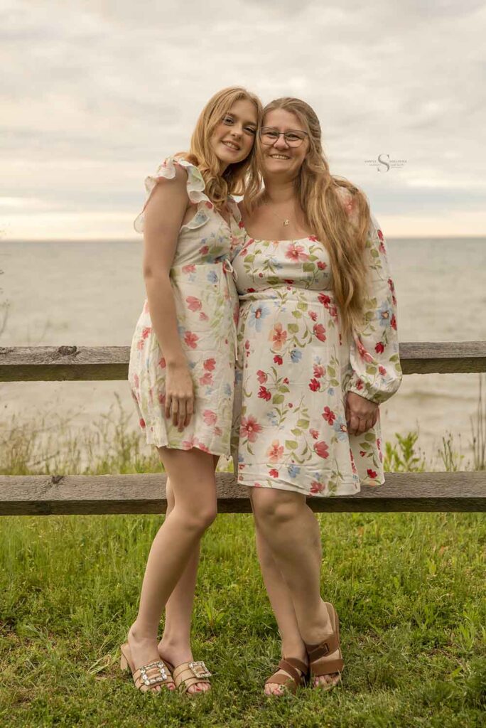 A mother and daughter , wearing matching floral gowns, pose for a portrait together in front of Lake Ontario at Mexico Point State Park with Simply Shelayna Family Photographer. 