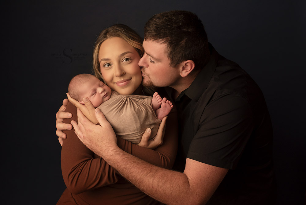 Father leans over and kisses mother on the cheek as she holds her swaddled newborn son for a family portrait with Simply Shelayna Newborn Photography in Syracuse NY.