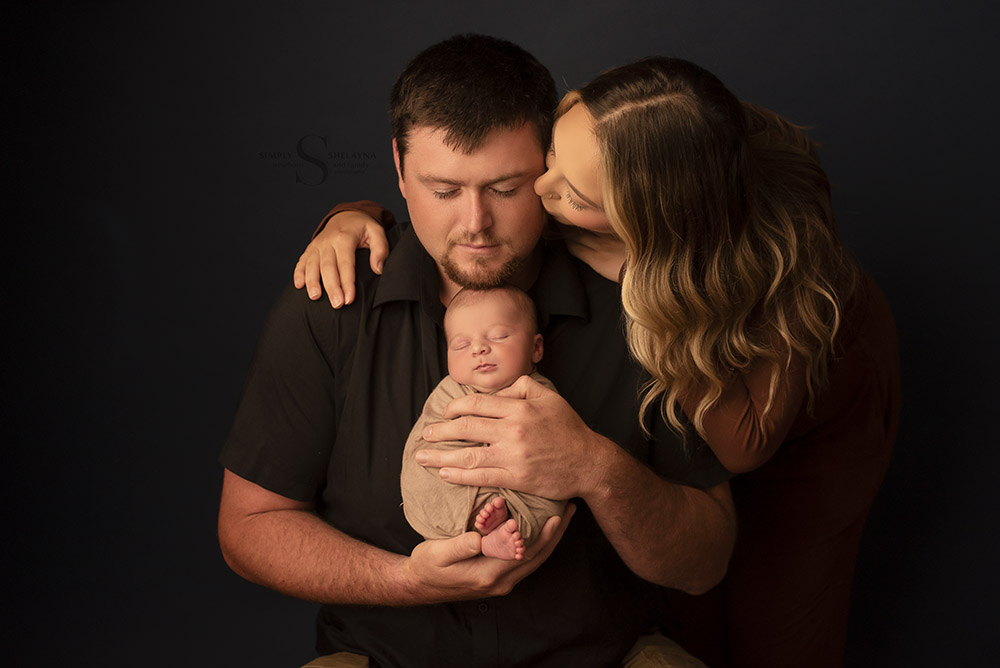 Mother leans over and kisses father on the cheek as he holds his swaddled newborn son for a family portrait with Simply Shelayna Newborn Photography in Syracuse NY.