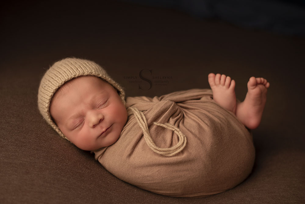 A baby boy is swaddled in a knit brown fabric with a hand knitted bonnet adorning his head for a newborn portrait with Simply Shelayna Family Photography in CNY.