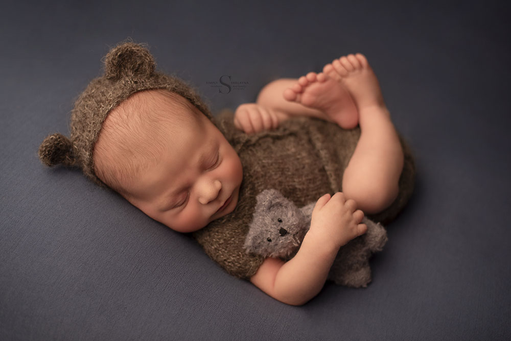 A newborn boy holds close a teddy bear as he sleeps with a bonnet and bear ears for a newborn portrait with Simply Shelayna Newborn Photography in Syracuse NY.