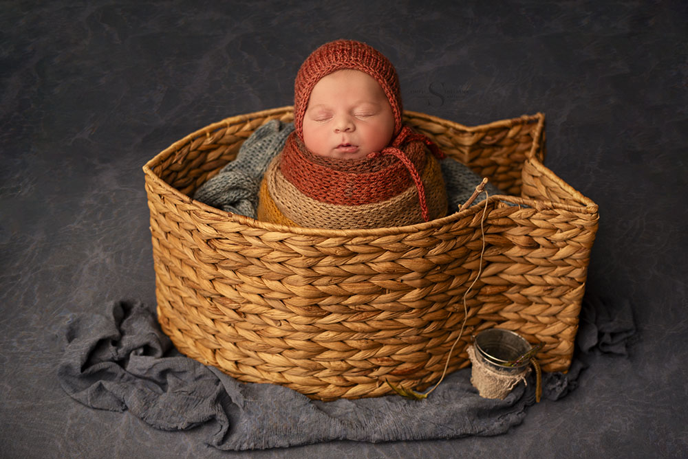 A baby boy is swaddled in potato sack pose in a hand knitted autumn colored wrap and placed in a fish shaped basket with Simply Shelayna Newborn Photography studio in Syracuse NY.