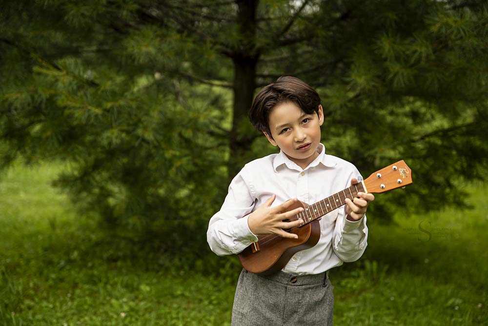 A young boy stands in front of an evergreen tree and plays his ukulele for a milestone portrait with Simply Shelayna Family photography in Syracuse NY. 