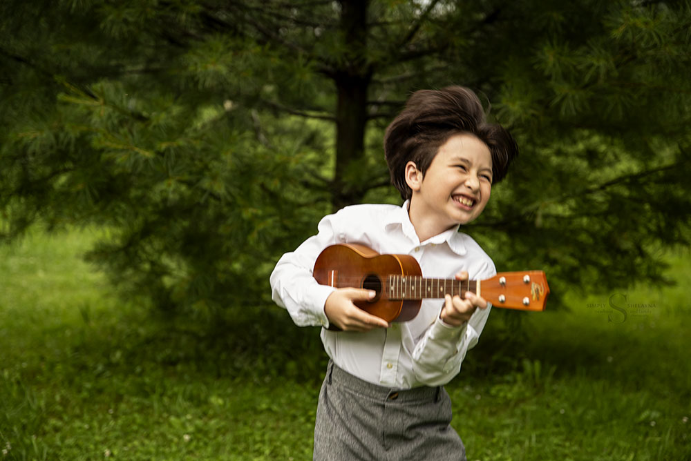 A young boy gleefully plays his ukulele while dressed in his orchestra attire for a  portrait with Simply Shelayna milestone Photography in CNY.