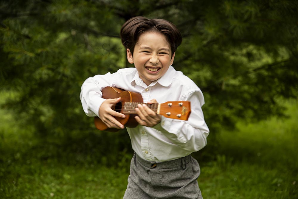 A young boy gleefully plays his ukulele while dressed in his orchestra attire for a  portrait with Simply Shelayna milestone Photography in CNY.