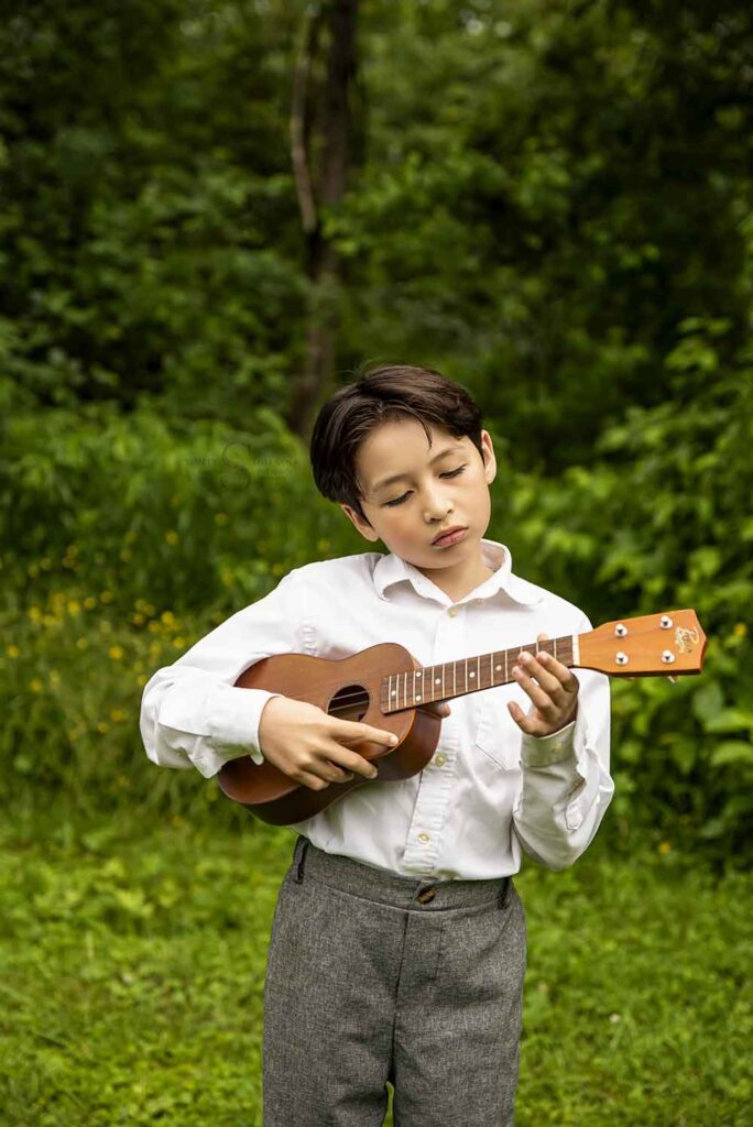 A young boy stands in front of an evergreen tree and plays his ukulele for a milestone portrait with Simply Shelayna Family photography in Syracuse NY. 